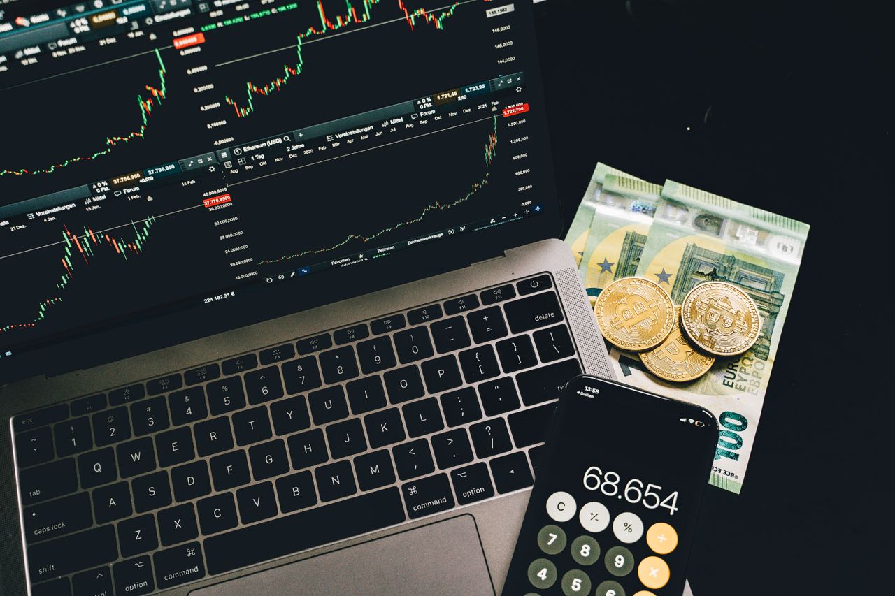 contact-img A laptop displaying stock charts with Bitcoin, Euros, and a cellphone calculator, showcasing financial analysis.